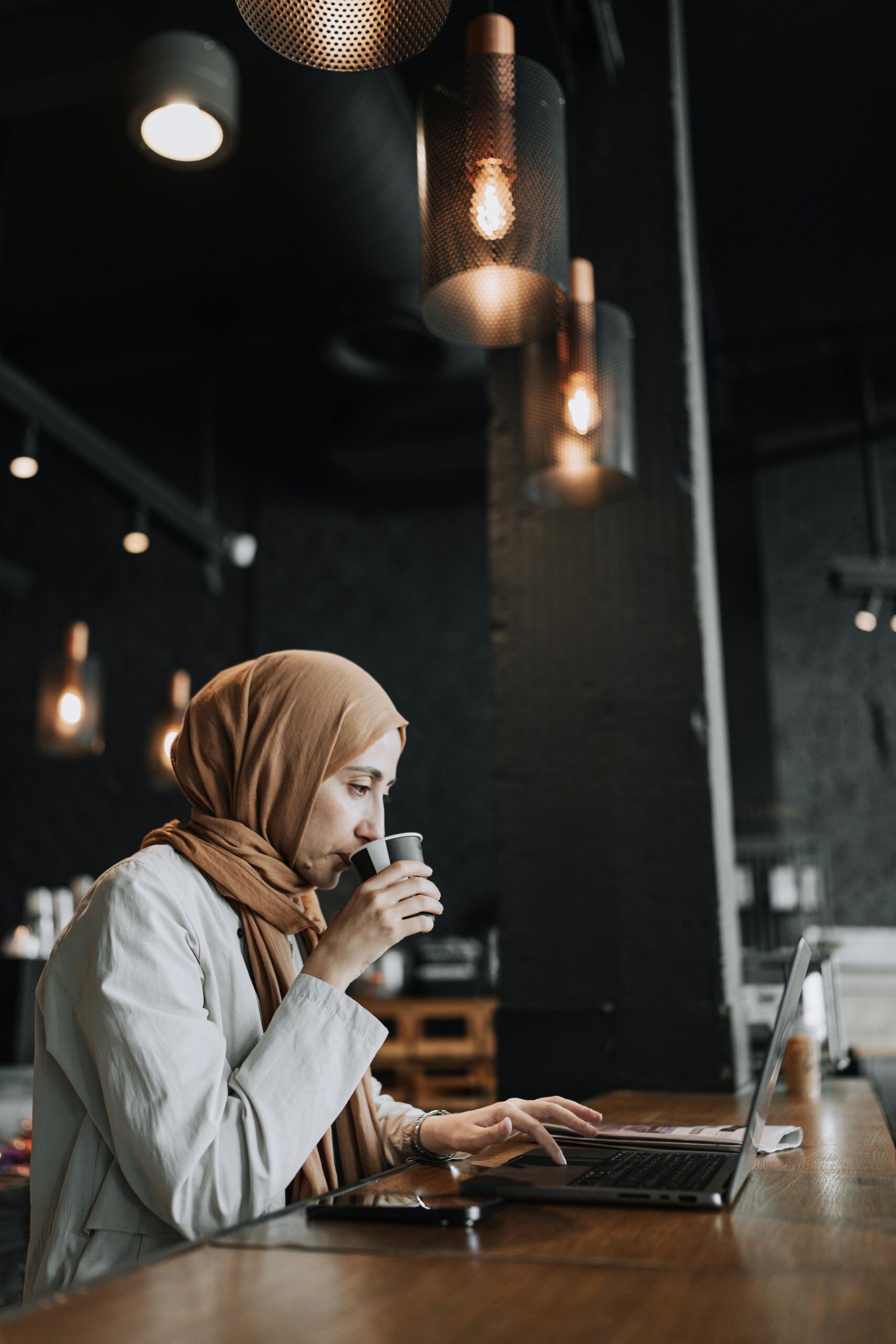 Woman in headscarf enjoys coffee while working on a laptop at a stylish café.